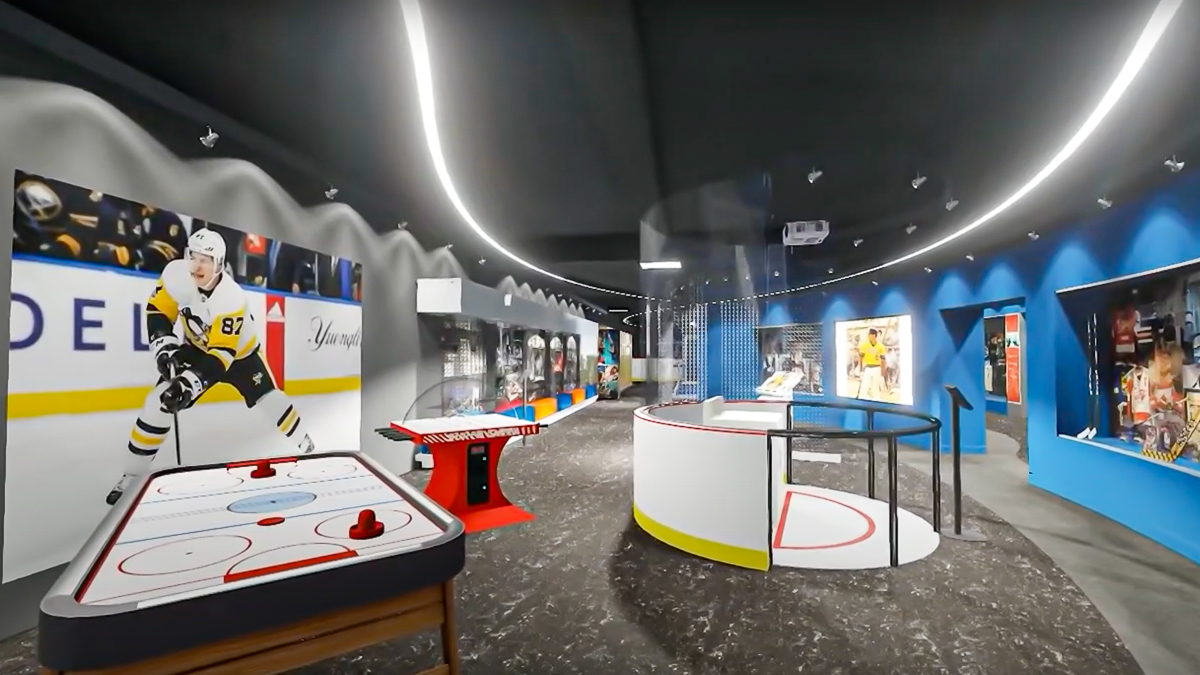 A virtual image of what the new Sydney Crosby exhibit will look like in the Sport Hall of Fame. There is three games set up in the middle of the room, air hockey, football and a mini ice rink. a picture of Sydney Crosby hangs on a grey wall. opposite of that is a blue wall with glass display cases featuring sport artifacts. the floor is dark grey with a black ceiling that has a long strip of lights running along it.